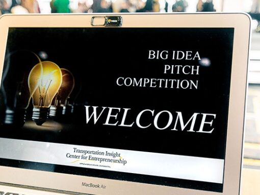 Laptop screen displaying Big Idea Pitch Competition welcome message with light bulb graphic in a meeting room.