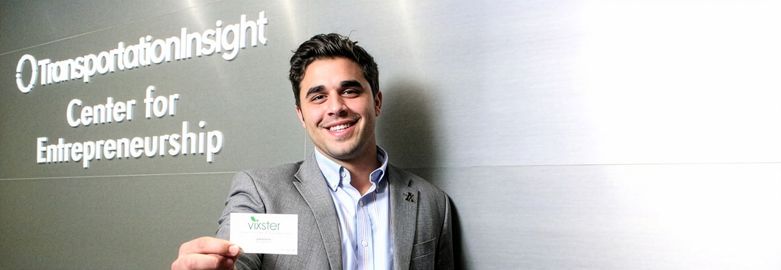 Mentor, Zak Ammar in business attire holding a card in front of a wall with Transportation Insight signage.